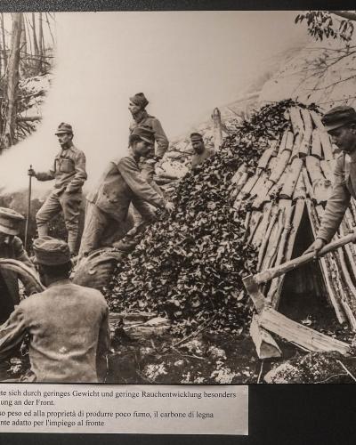 A black and white historical photograph showing soldiers in World War I-era uniforms constructing a charcoal kiln in a forest, with a caption below in German and Italian explaining the utility of charcoal for the front lines.