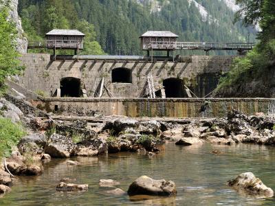 A stone dam structure with two wooden control houses on top, situated in a forested mountain valley with a stream flowing over rocks in the foreground.