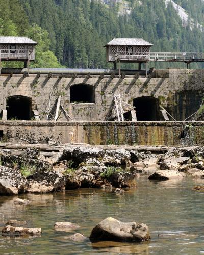A stone dam structure with two wooden control houses on top, situated in a forested mountain valley with a stream flowing over rocks in the foreground.