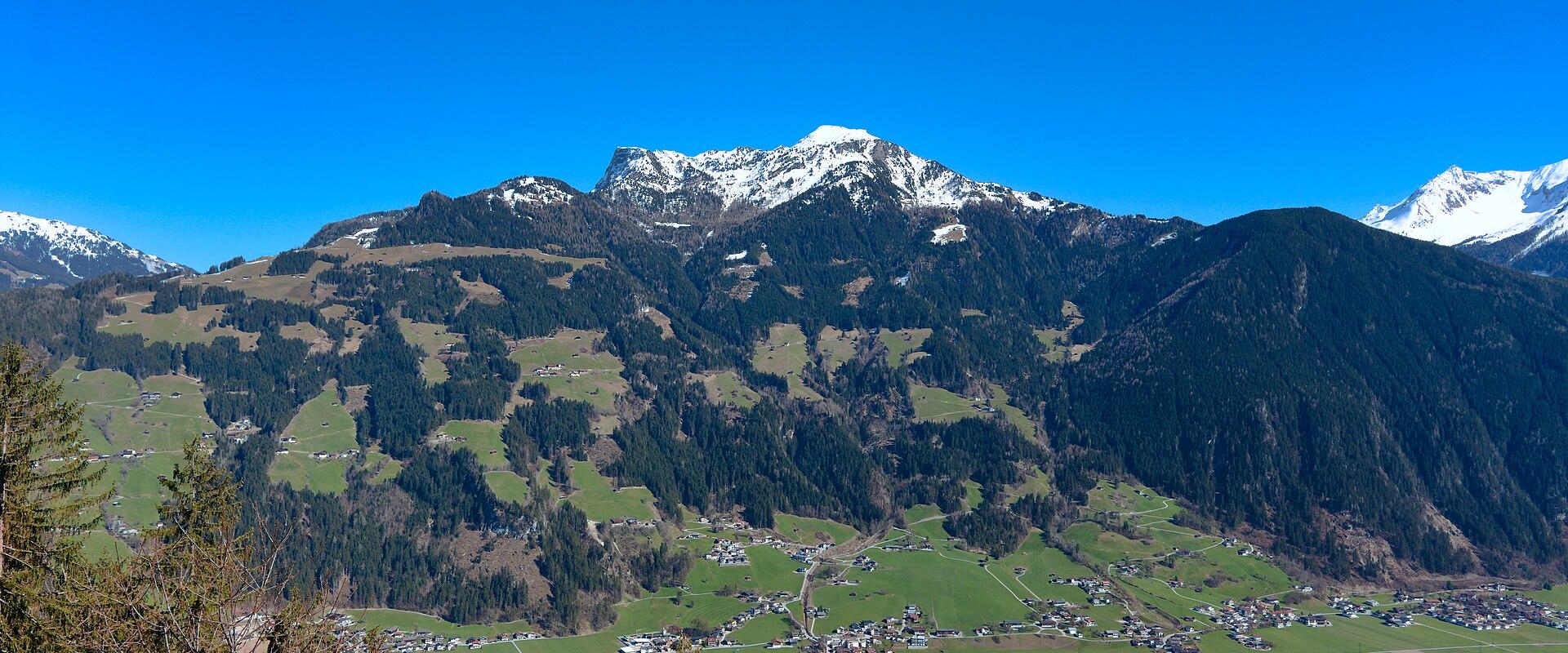 Snow-capped mountains rise above a lush green valley dotted with small buildings, under a clear blue sky.