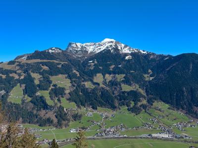 Snow-capped mountains rise above a lush green valley dotted with small buildings, under a clear blue sky.