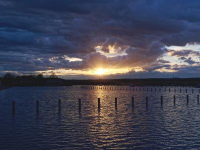 A sunset view over a body of water with rows of wooden pilings in the foreground and a distant shoreline with trees and buildings.