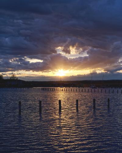 A sunset view over a body of water with rows of wooden pilings in the foreground and a distant shoreline with trees and buildings.