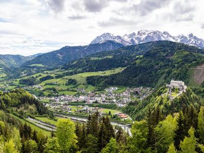 A panoramic landscape view of a valley town with a prominent white fortress perched on a hillside, surrounded by green slopes and distant snow-capped mountains.