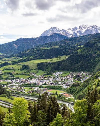 A panoramic landscape view of a valley town with a prominent white fortress perched on a hillside, surrounded by green slopes and distant snow-capped mountains.