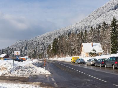 A snowy roadside scene on a mountain pass featuring a wet asphalt road, parked cars, a yellow van, a white house with a snow-covered roof, and a snow-dusted forested hillside in the background; a signpost with a phone number and a blue parking symbol