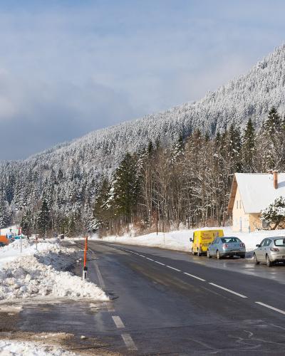 A snowy roadside scene on a mountain pass featuring a wet asphalt road, parked cars, a yellow van, a white house with a snow-covered roof, and a snow-dusted forested hillside in the background; a signpost with a phone number and a blue parking symbol