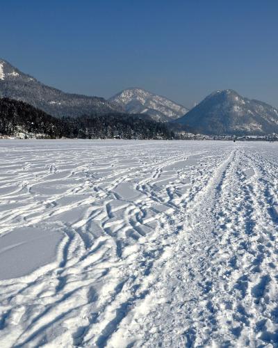A wide, snow-covered frozen lake surface with numerous tracks, backed by forested hills and snow-capped mountains under a clear blue sky.