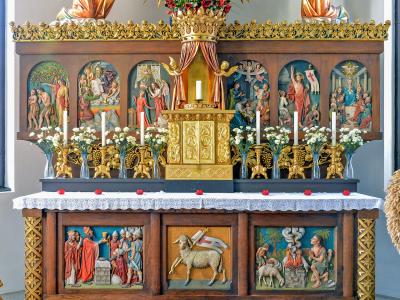 A close-up view of an ornate wooden church altar featuring a central golden tabernacle, flanked by painted arched panels depicting religious scenes, and lower relief carvings including a lamb with a flag.