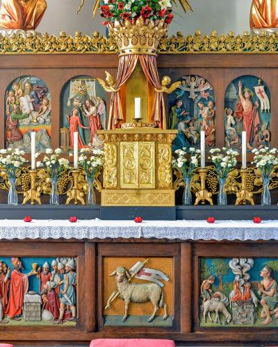 A close-up view of an ornate wooden church altar featuring a central golden tabernacle, flanked by painted arched panels depicting religious scenes, and lower relief carvings including a lamb with a flag.