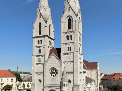 A high-angle photograph of a large, light-colored stone cathedral with two tall, pointed spires, situated in a paved town square with a parking lot and playground nearby.