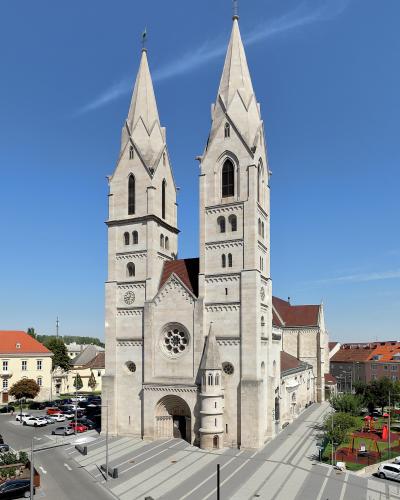 A high-angle photograph of a large, light-colored stone cathedral with two tall, pointed spires, situated in a paved town square with a parking lot and playground nearby.