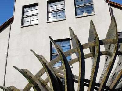 A close-up view of a large, weathered wooden water wheel structure positioned in front of a white, multi-story building with rectangular windows under a blue sky.