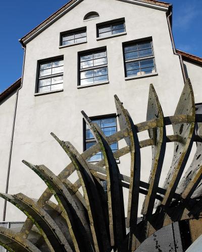 A close-up view of a large, weathered wooden water wheel structure positioned in front of a white, multi-story building with rectangular windows under a blue sky.