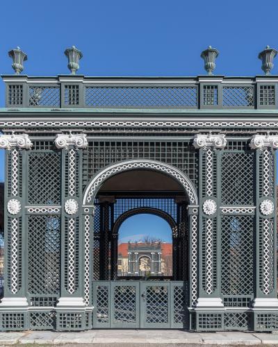 A green and white ornate ironwork gate with classical columns and decorative lattice, set against a blue sky with bare trees on either side.