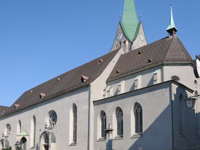 A large, light-colored Gothic-style church with a prominent green spire and clock tower, situated in a paved square with a fountain in the foreground.