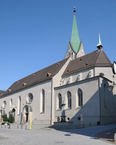 A large, light-colored Gothic-style church with a prominent green spire and clock tower, situated in a paved square with a fountain in the foreground.