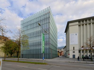 A modern glass cube building (Kunsthaus Bregenz) next to a concrete building, with a green vertical banner reading "Jeff Wall" and a white poster on the adjacent building.