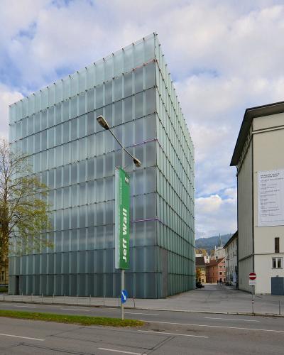 A modern glass cube building (Kunsthaus Bregenz) next to a concrete building, with a green vertical banner reading "Jeff Wall" and a white poster on the adjacent building.