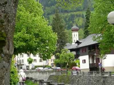 A paved walkway alongside a river in a town, with traditional buildings and a church tower visible in the background.