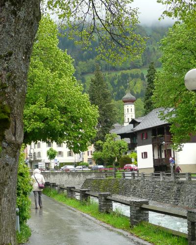 A paved walkway alongside a river in a town, with traditional buildings and a church tower visible in the background.