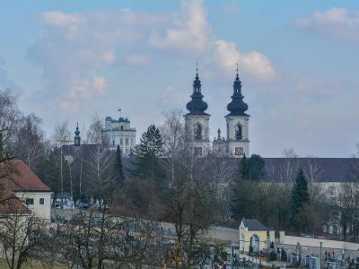 A view of a large white monastery complex with two prominent dark onion-domed towers rising above a cemetery in the foreground, with a white building flying a flag visible to the left.