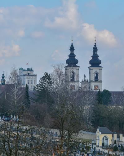 A view of a large white monastery complex with two prominent dark onion-domed towers rising above a cemetery in the foreground, with a white building flying a flag visible to the left.