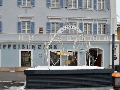 A large white metal sculpture of bicycle wheels and a sign reading "RADSTADT" and "RADGARTEN" situated on a snow-covered base in front of a light blue building with "RAIFFEISENBANK" signage.