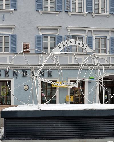A large white metal sculpture of bicycle wheels and a sign reading "RADSTADT" and "RADGARTEN" situated on a snow-covered base in front of a light blue building with "RAIFFEISENBANK" signage.