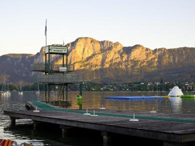 A wooden pier extending into a lake with a tall green lifeguard tower, mountains in the background, and floating water play structures.