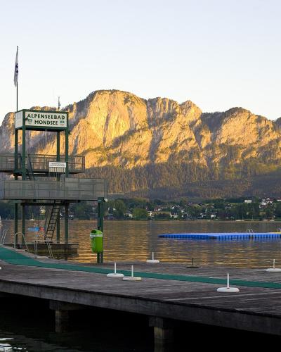 A wooden pier extending into a lake with a tall green lifeguard tower, mountains in the background, and floating water play structures.