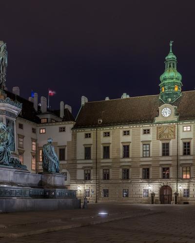 A night photograph of a large bronze equestrian-style monument featuring a standing central figure and seated allegorical figures on a pedestal, situated in a paved courtyard in front of a long, illuminated palace building with a distinctive green co