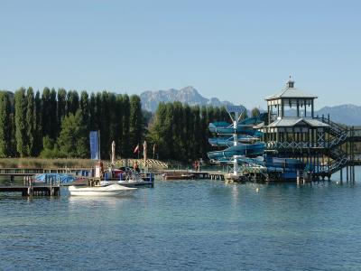 A large wooden water slide tower with blue slides and a gazebo-style roof structure situated on a lake, with mountains and trees in the background.