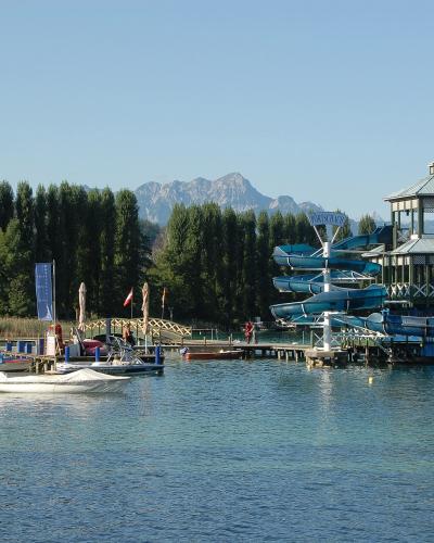 A large wooden water slide tower with blue slides and a gazebo-style roof structure situated on a lake, with mountains and trees in the background.