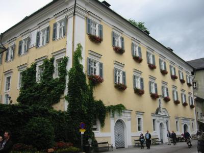 A large, pale yellow building with grey shutters, flower boxes, and ivy climbing the facade, situated on a street corner with pedestrians and a traffic sign visible.