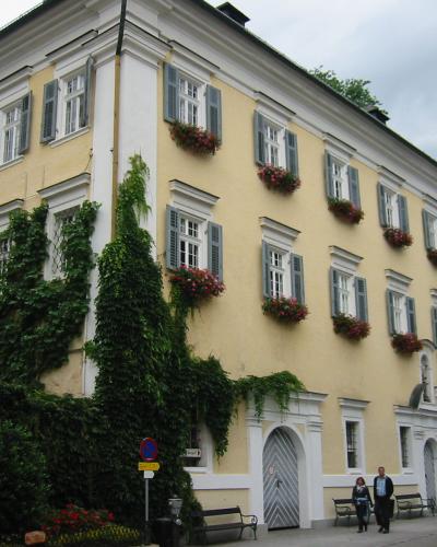 A large, pale yellow building with grey shutters, flower boxes, and ivy climbing the facade, situated on a street corner with pedestrians and a traffic sign visible.
