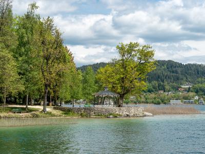 A scenic view of a lake shoreline featuring a stone-walled peninsula with a gazebo, surrounded by trees and reeds, with a forested hill and buildings in the background.