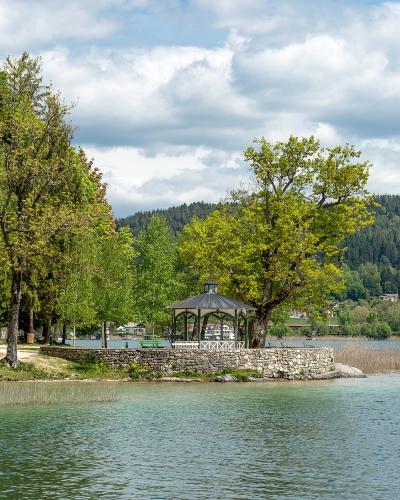 A scenic view of a lake shoreline featuring a stone-walled peninsula with a gazebo, surrounded by trees and reeds, with a forested hill and buildings in the background.