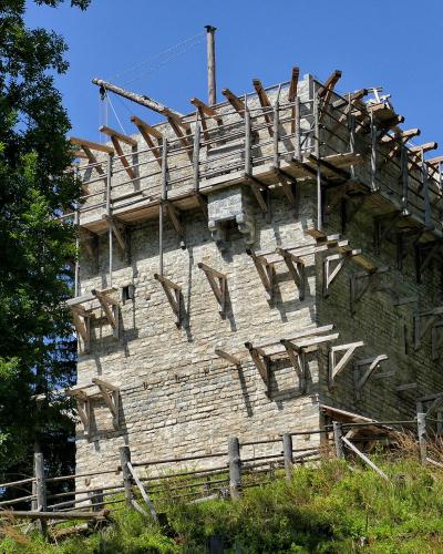 A stone tower under construction with extensive wooden scaffolding and support beams, situated on a grassy hill surrounded by trees.