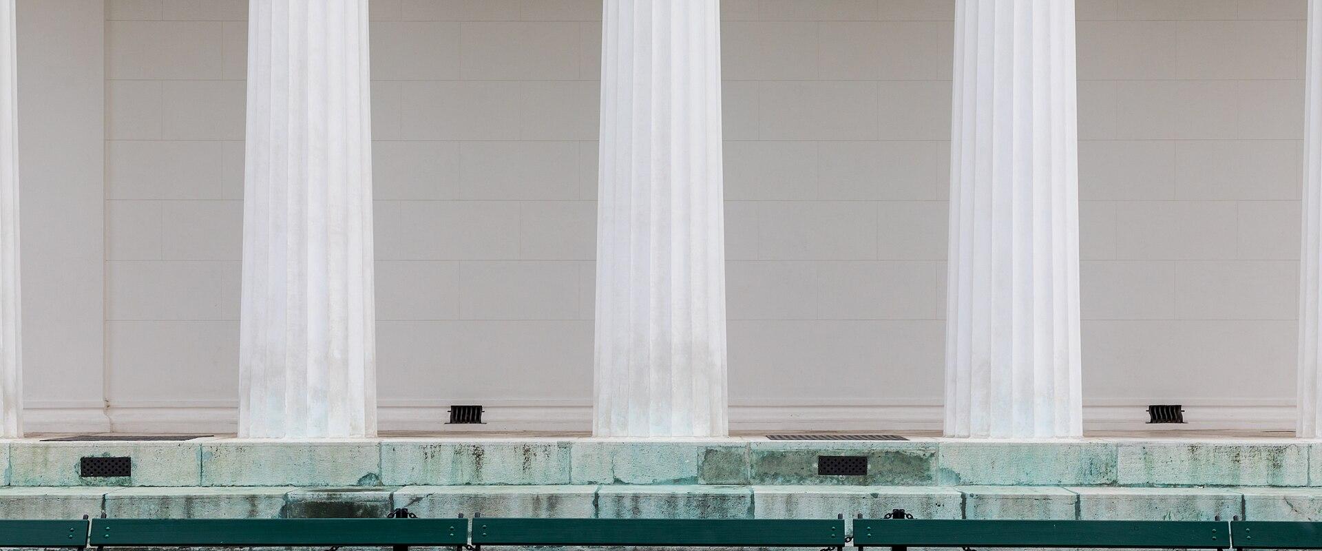 The image shows the base and lower columns of a classical-style temple with white fluted columns, a stone platform, and green park benches in the foreground.
