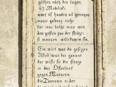 A close-up photograph of a rectangular stone plaque mounted on a textured exterior wall, featuring two stanzas of medieval German text in black calligraphy and a citation at the bottom reading "Nibelungenlied St. 1268...1269".