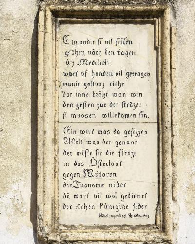 A close-up photograph of a rectangular stone plaque mounted on a textured exterior wall, featuring two stanzas of medieval German text in black calligraphy and a citation at the bottom reading "Nibelungenlied St. 1268...1269".
