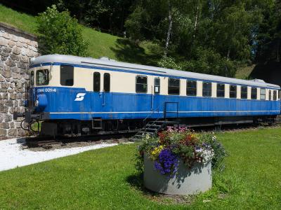 A blue and cream-colored ÖBB railcar (number 5144 001-4) is parked on a short section of track next to a stone retaining wall, with a flower planter in the foreground and a grassy hillside behind it.