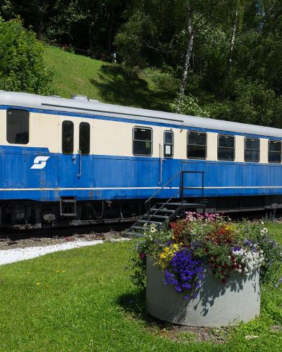 A blue and cream-colored ÖBB railcar (number 5144 001-4) is parked on a short section of track next to a stone retaining wall, with a flower planter in the foreground and a grassy hillside behind it.