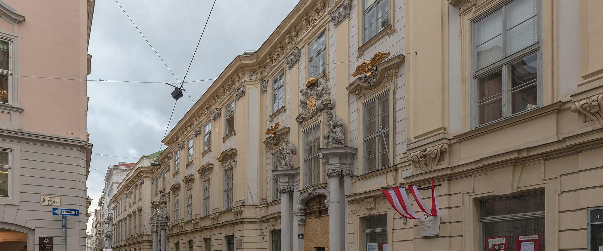 The image shows a long, ornate Baroque-style building with a cream facade, decorative pilasters, sculptural elements including eagles and coats of arms, and Austrian flags hanging from the ground floor; street signs indicate “Jägerstraße” and “Kär