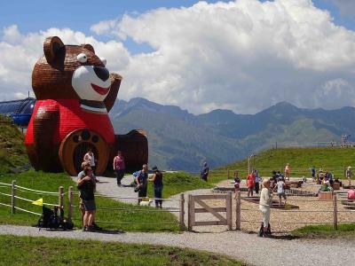A large wooden bear sculpture wearing a red sweater stands on a grassy hillside next to a playground area with people, set against a backdrop of green mountains under a cloudy sky.