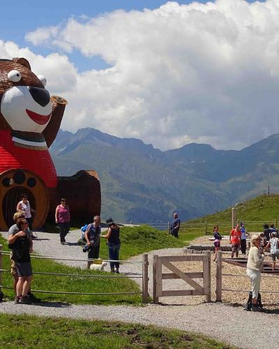 A large wooden bear sculpture wearing a red sweater stands on a grassy hillside next to a playground area with people, set against a backdrop of green mountains under a cloudy sky.