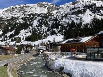 A river flows through a village with traditional alpine buildings, backed by a steep, snow-dusted mountain slope featuring distinct horizontal terracing patterns, under a blue sky with a single cloud.