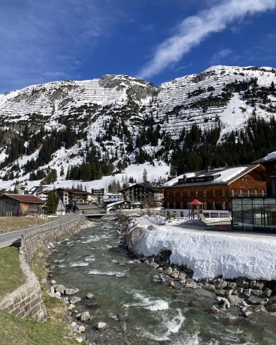 A river flows through a village with traditional alpine buildings, backed by a steep, snow-dusted mountain slope featuring distinct horizontal terracing patterns, under a blue sky with a single cloud.