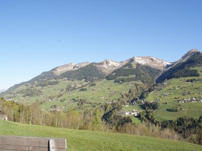 A scenic view of a green alpine valley with scattered houses and a church, looking towards snow-dusted mountain peaks under a clear blue sky, with a wooden bench in the foreground.